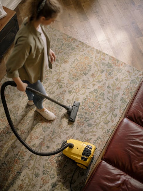 A woman vacuuming a patterned area rug in a living room with wooden flooring and a brown leather sofa. She is using a yellow vacuum cleaner with a black hose, focusing on the central part of the rug to remove dust and dirt. The room is well-lit, showcasing the textured, multi-colored floral design of the rug, which appears clean and well-maintained. The scene emphasizes surface cleaning and deep cleaning practices, with Barnet Carpet Cleaning experienced in residential carpet and rug sanitisation. The setting reflects a tidy, hygienic domestic space prepared for regular maintenance or professional cleaning services provided by Barnet Carpet Cleaning.