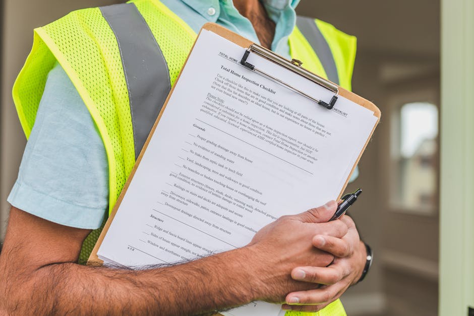 Close-up of a person wearing a high-visibility yellow safety vest and light blue shirt, holding a clipboard with a detailed 'Total Home Inspection Checklist' document. The individual is securing the clipboard with their left hand and writing with a black pen in their right hand. The background shows a softly lit indoor space with light-colored walls and a window, indicating a residential or commercial property setting. The scene emphasizes thorough surface cleaning, sanitisation, and inspection processes typical of end of tenancy cleaning services provided by Barnet Carpet Cleaning, including tools like the clipboard and pen used for documentation in a clean, organized environment.