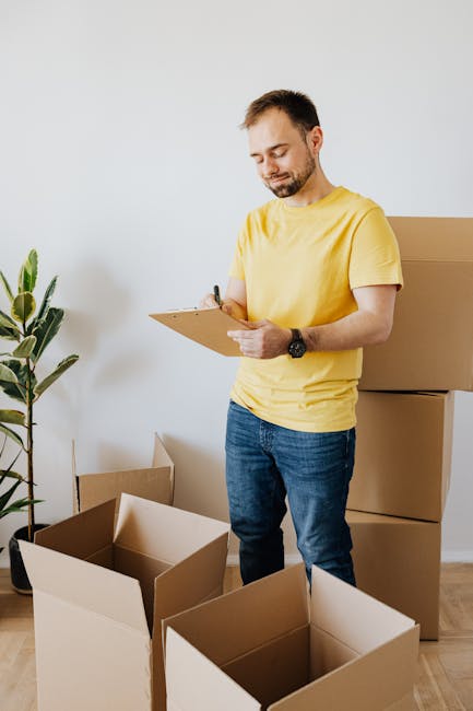 Close-up of a person wearing a high-visibility yellow safety vest and light blue shirt, holding a clipboard with a detailed 'Total Home Inspection Checklist' document. The individual is securing the clipboard with their left hand and writing with a black pen in their right hand. The background shows a softly lit indoor space with light-colored walls and a window, indicating a residential or commercial property setting. The scene emphasizes thorough surface cleaning, sanitisation, and inspection processes typical of end of tenancy cleaning services provided by Barnet Carpet Cleaning, including tools like the clipboard and pen used for documentation in a clean, organized environment.