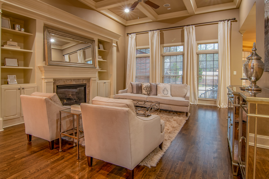A bright, well-lit living room featuring a beige vaulted ceiling with recessed lighting. The focal point is a fireplace with a beige mantel and a large rectangular mirror above it, reflected in built-in shelves on either side containing decorative items. The space includes a light-colored upholstered sofa with patterned cushions, positioned near large windows draped with cream curtains. Two matching armchairs are arranged around a glass-topped coffee table on a plush, cream area rug. The room has polished wooden flooring, with a sideboard on the right decorated with a large metallic vase. Natural light filters through the windows, illuminating the tidy, hygienic surface of the furniture and floors. Barnet Carpet Cleaning specializes in deep cleaning and sanitisation of domestic spaces, helping maintain cleanliness and hygiene in homes like this one.