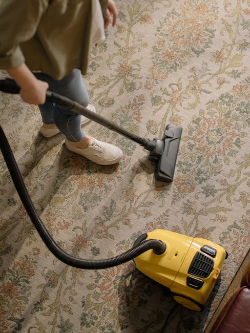 A person using a vacuum cleaner with a yellow body and black hose to deep clean a patterned oriental-style carpet in a residential living room. The individual, wearing beige sneakers and jeans, is holding the vacuum's handle while moving it across the surface. The room features a timber floor visible around the carpet, and soft, natural lighting enhances the cleanliness and condition of the rug. The scene depicts a routine domestic cleaning process, highlighting surface cleaning and maintenance, with the equipment supplied by Barnet Carpet Cleaning for comprehensive hygiene and care of home flooring surfaces.