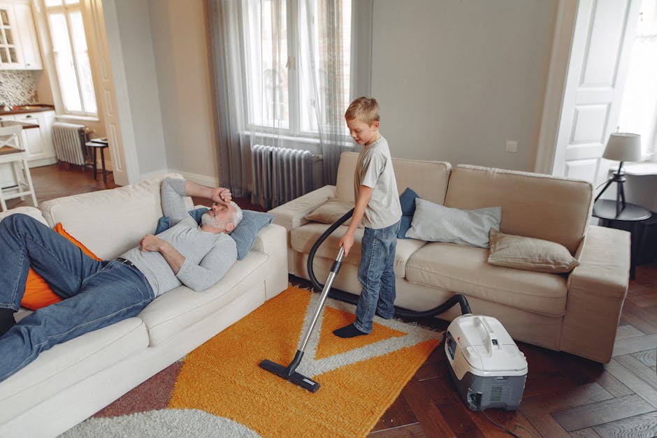 A young boy using a vacuum cleaner to thoroughly clean the beige carpet in a living room, with an elderly man lying comfortably on a white sofa nearby. The room features light grey walls, large windows with sheer curtains allowing natural light, and wooden flooring partially covered by a multicolored area rug. The space is furnished with a beige couch with several cushions, a side table with a lamp, and a radiator below the window. Visible cleaning equipment includes a modern portable vacuum cleaner, and the entire scene depicts a deep cleaning process aimed at maintaining hygiene and freshness in a domestic setting, with Barnet Carpet Cleaning providing professional service for surface cleaning and sanitisation.