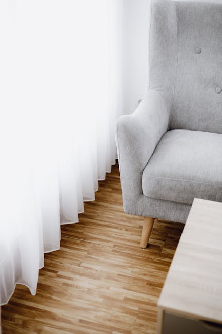 Close-up view of a light grey upholstered armchair positioned next to a window with sheer white curtains, in a residential living room. The armchair features a soft fabric surface, with visible tufted buttons on the backrest, and wooden tapered legs. The wooden floor has a natural finish, exhibiting a smooth, clean, and well-maintained surface. A small light-colored wooden table is partially visible in the foreground. The lighting is natural, coming through the curtains, highlighting the cleanliness and freshness of the room. Barnet Carpet Cleaning offers professional surface cleaning and sanitisation services for homes, ensuring hygiene and deep cleaning of furniture and interior spaces.