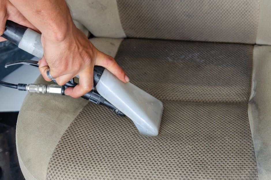 A close-up of a person's hand holding a handheld steam cleaning device, which is pressed against a beige fabric-upholstered car seat. The device emits steam to deep clean the fabric, visibly lifting dirt and stains from the textured surface. The seat has a combination of plain and patterned fabric, both showing signs of previous use. The setting appears to be in a vehicle interior, with soft lighting highlighting the cleanliness process. Barnet Carpet Cleaning employs professional surface cleaning techniques to ensure domestic and vehicle upholstery is hygienically maintained, emphasizing thorough sanitisation and effective removal of dust and dirt from fabric surfaces.